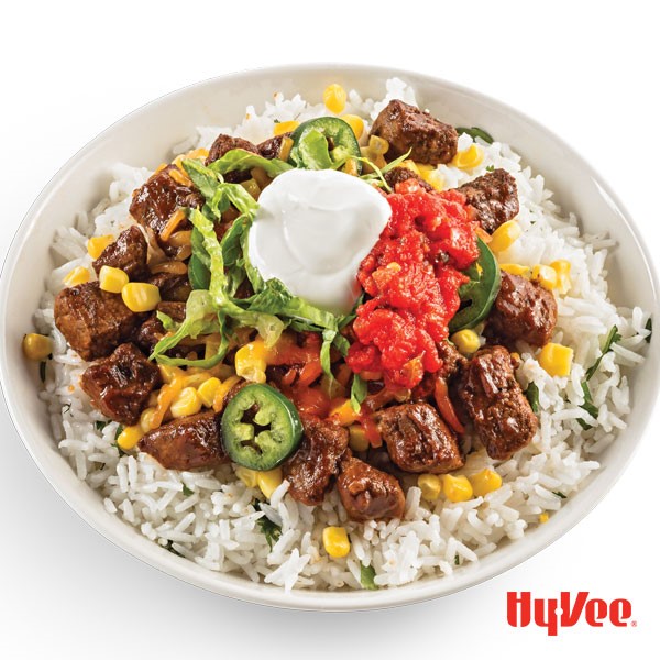 White bowl filled with cilantro rice, beef, sliced jalapenos, corn kernels, red salsa, and sour cream