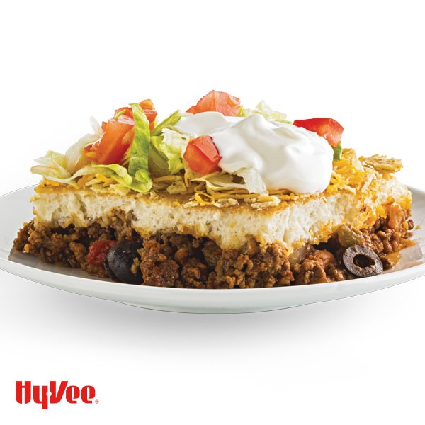 Layered taco bake with ground beef, sliced olives, dough, shredded cheese, shredded lettuce and a dollop of sour cream
