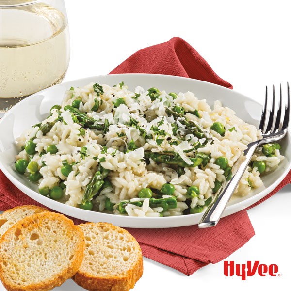 Shallow dish filled with risotto, peas, asparagus, and grated Parmesan cheese with a side of sliced bread, white wine, and fork
