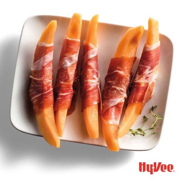 Cantalope wedges wrapped in prosciutto on a white plate with thyme sprigs