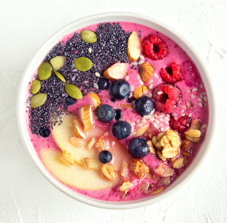 Pink smoothie served in a white bowl, topped with fresh fruit, nuts, seeds and granola
