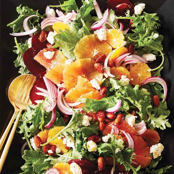 Black platter with gold silverware holding salad with sugared almonds, crumbled goat cheese, sliced red onions, and whole slices of blood oranges