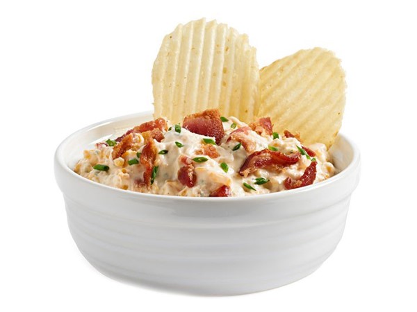 White bowl filled with dip and topped with cooked chopped  bacon, chopped chives, and two crinkled potato chips