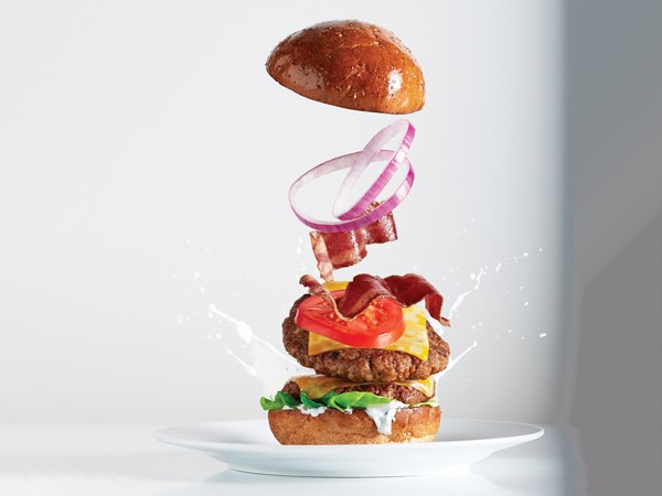 Burger patty, toppings and top bun in mid-air above a white plate 